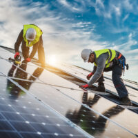 Handwerker auf einem Wohnhausdach beim Bau einer Photovoltaikanlage (PV-Anlage). Zwei Arbeiter auf der linken Seite des Daches tragen Sicherheitsausrüstung und montieren Solarmodule auf einer modernen Dachfläche.