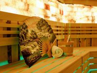 Ein großer bedruckter Saunafächer, ein gerolltes Handtuch und ein Sauna-Eimer in einer mit LEDs beleuchteten Saunakabine dekorativ hingestellt.