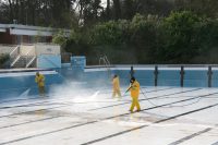 In gelbe Schutzanzüge gekleidete Personen reinigen ein leeres Schwimmbecken mit Hochdruckreinigern.