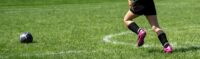 Die Beine eines Fußball spielenden Kindes mit pinken Turnschuhen auf einem grünen Fußballfeld.